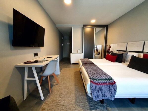 A modern hotel room with a flat-screen TV, a desk with a chair, a bed with a knitted blanket, and a large mirror.