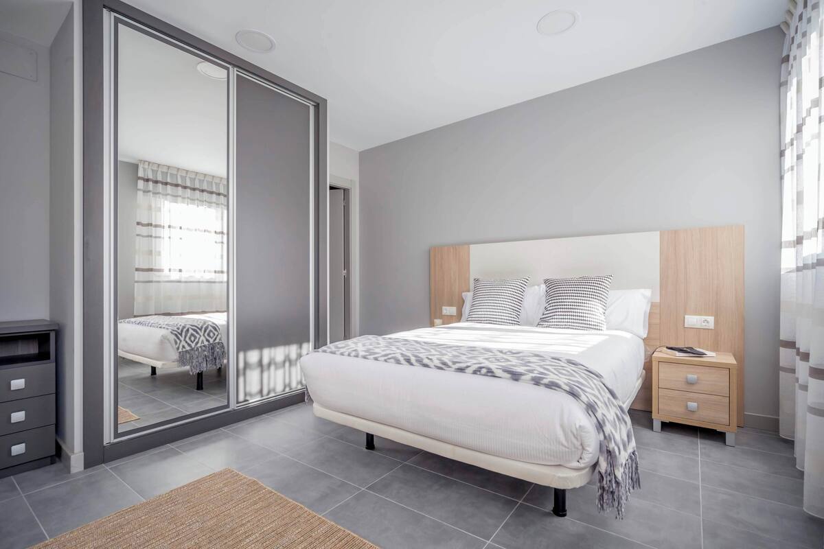A modern bedroom with a large bed, a wardrobe with a mirror, a bedside table, and a nightstand.