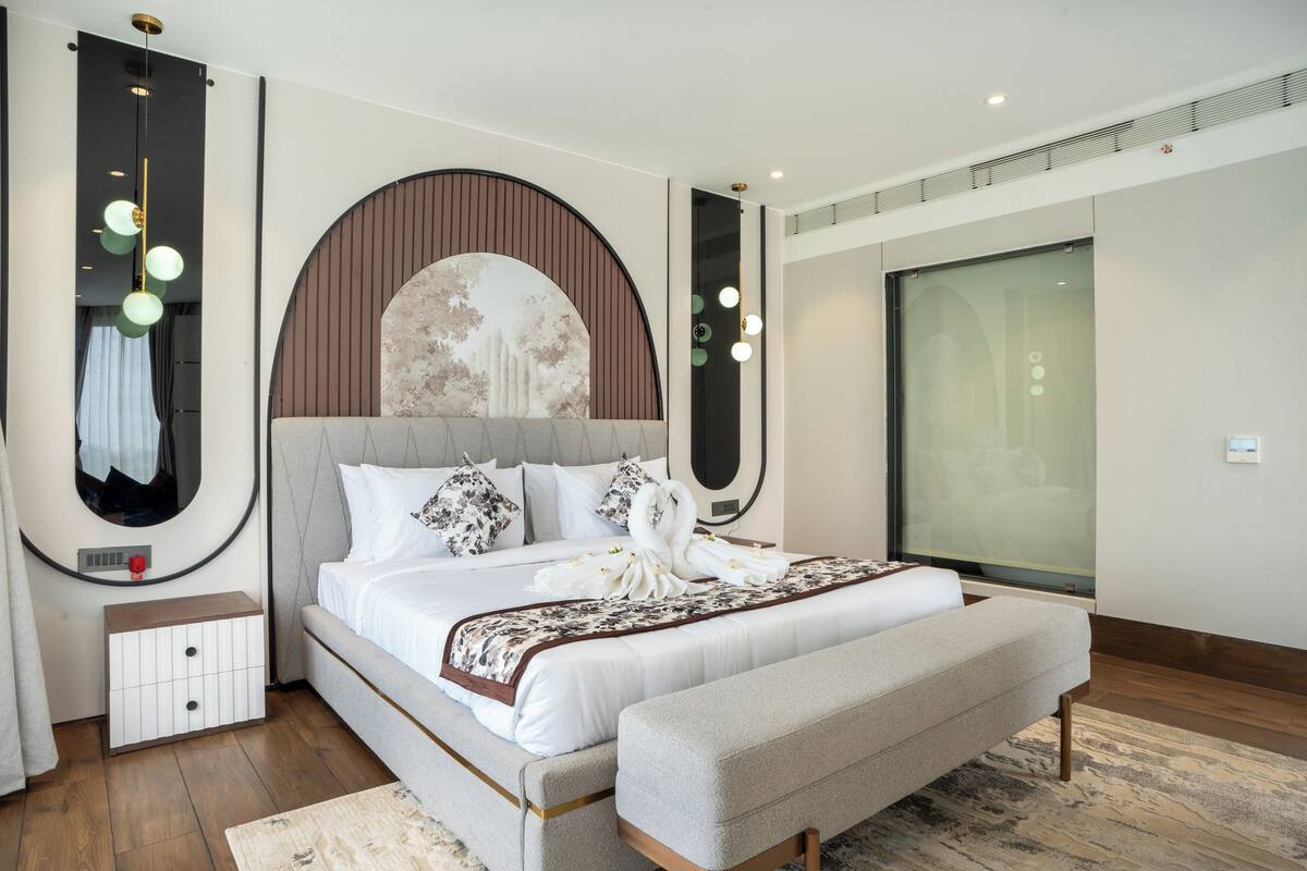 A modern hotel room with a large bed, a bench, a wall-mounted artwork, and a large window.