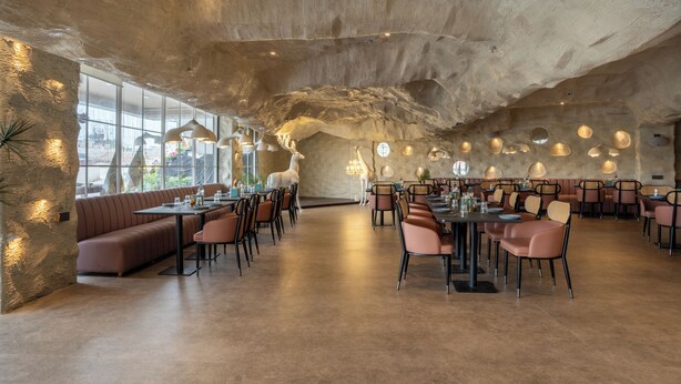 Een ruim restaurant met hoge plafonds, een centrale zithoek en uitzicht door grote ramen.
