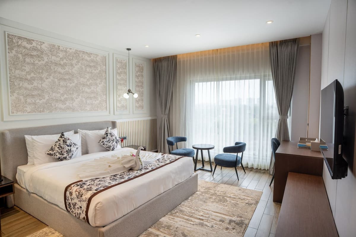 A hotel room with a large bed, a desk, two chairs, a TV, and a window with curtains.