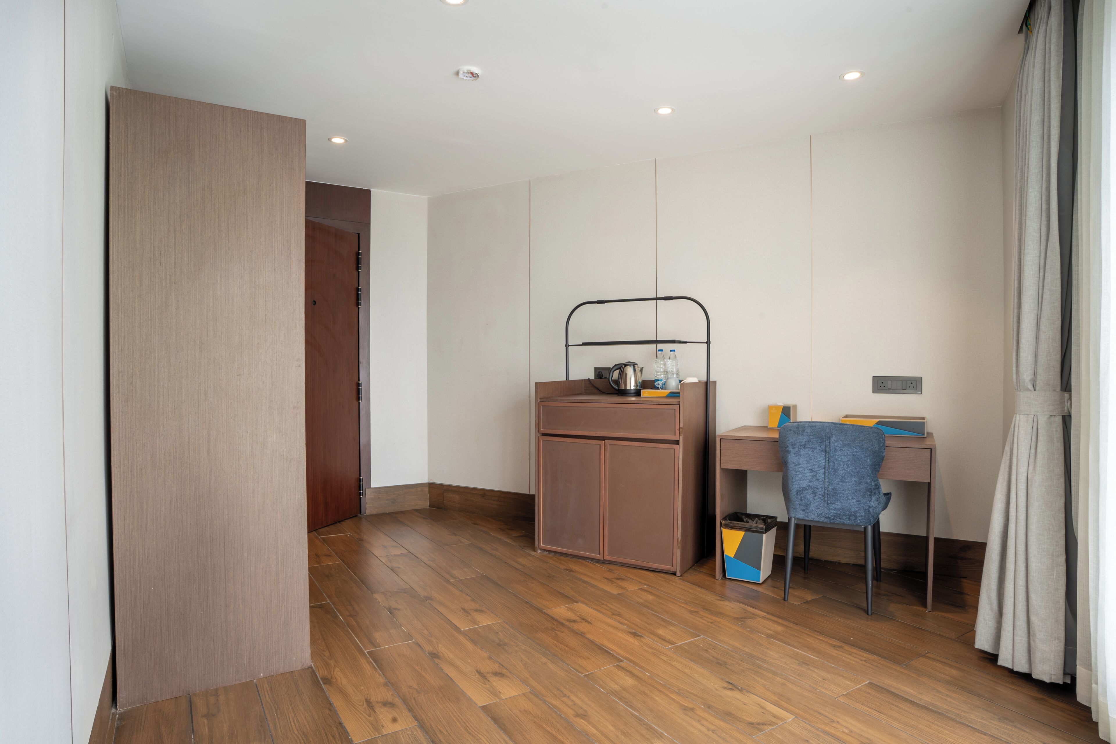 Una habitación con piso de madera, un escritorio con una silla, un mueble pequeño y una repisa metálica con una tetera.