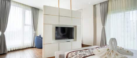 A modern hotel room with a large bed, a flat-screen TV mounted on a wall, a sofa, and a large window with curtains.