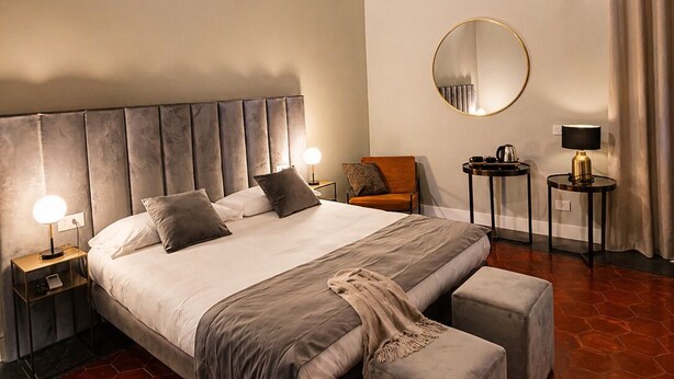 A hotel room with a large bed, a round mirror, two bedside tables with lamps, a small round table with a lamp, and a large window with curtains. - Cuevas Home Relais  (Naples)