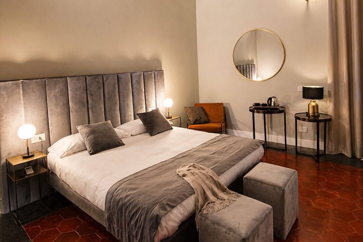 A hotel room with a large bed, a round mirror, two bedside tables with lamps, a small round table with a lamp, and a large window with curtains.