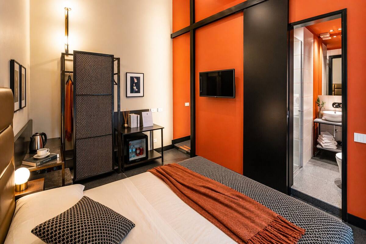 A hotel room with a bed, a desk with a lamp, a wardrobe, a TV, and a bathroom visible through an open door.