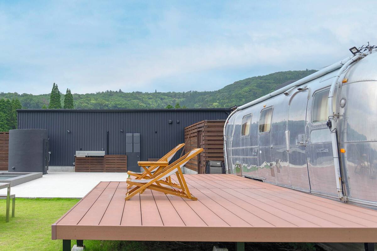 A modern RV with a transparent side, a wooden deck with benches, and a view of a green landscape.