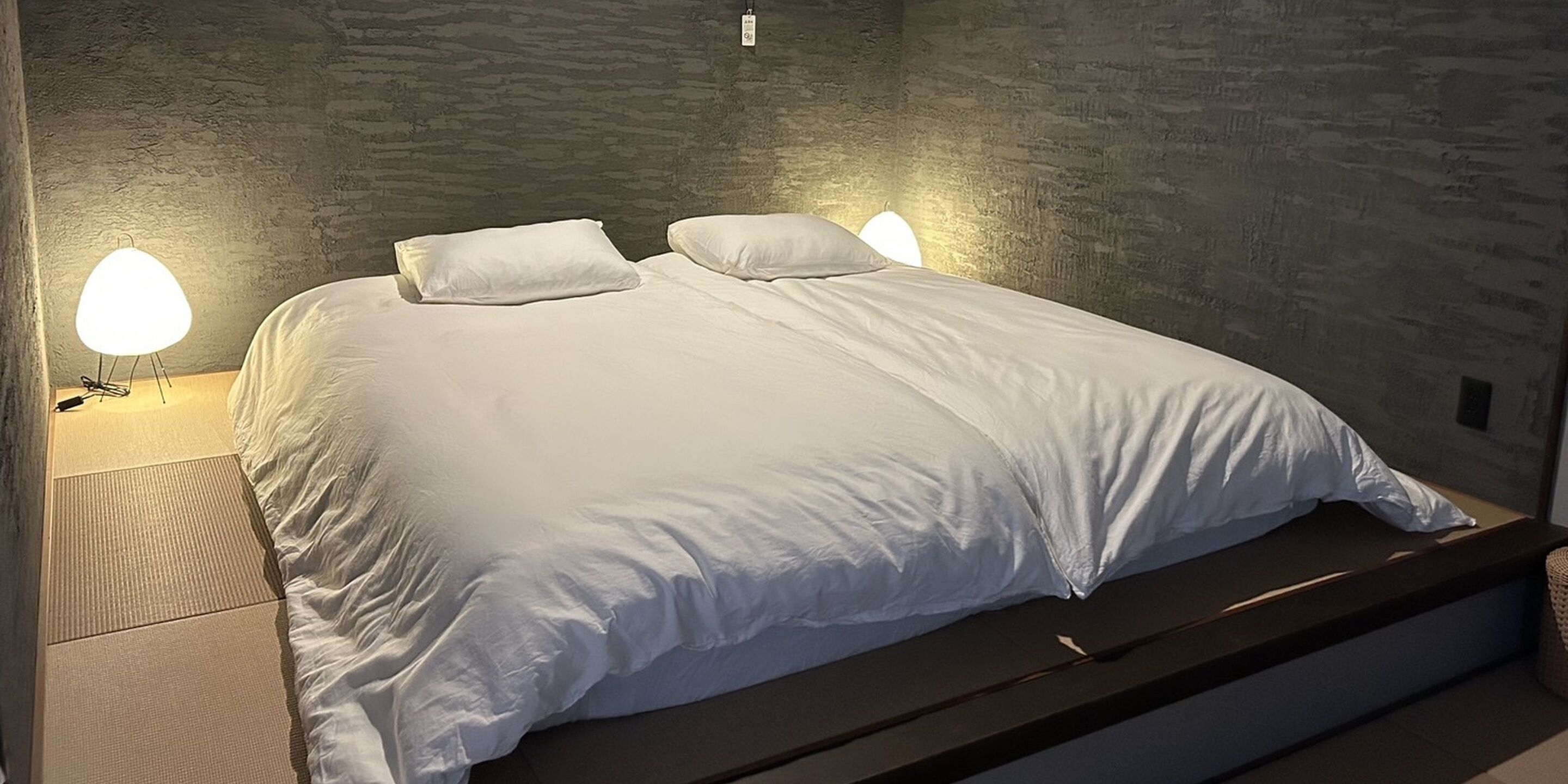 Ein ordentlich bezogenes Bett mit Kissen, einer Nachttischlampe und einer strukturierten Wand.
