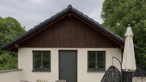 A modern house with a dark wooden facade, a grey door, and a balcony with outdoor furniture including a table, chairs, and a hanging chair.