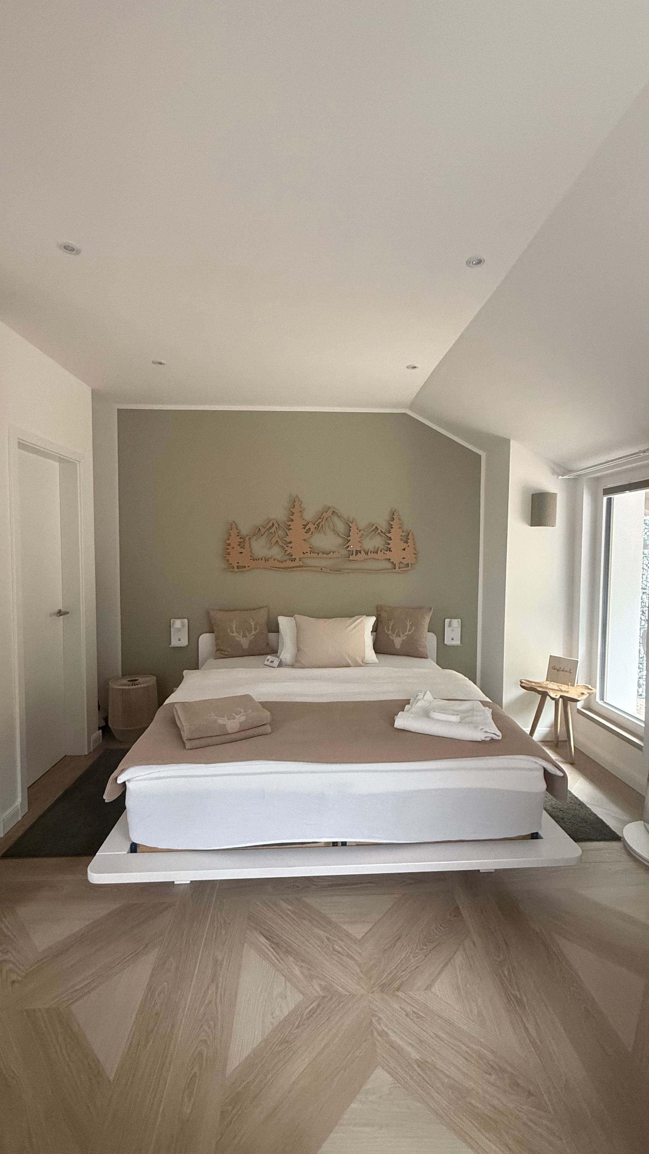 A modern bedroom with a large bed, a wall-mounted forest-themed decoration, and a window providing natural light.