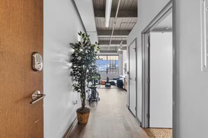 Un couloir avec un pot de plante, une porte dotée d’un clavier pour l’ouverture à code, et une vue sur une pièce contenant un bureau et une chaise.
