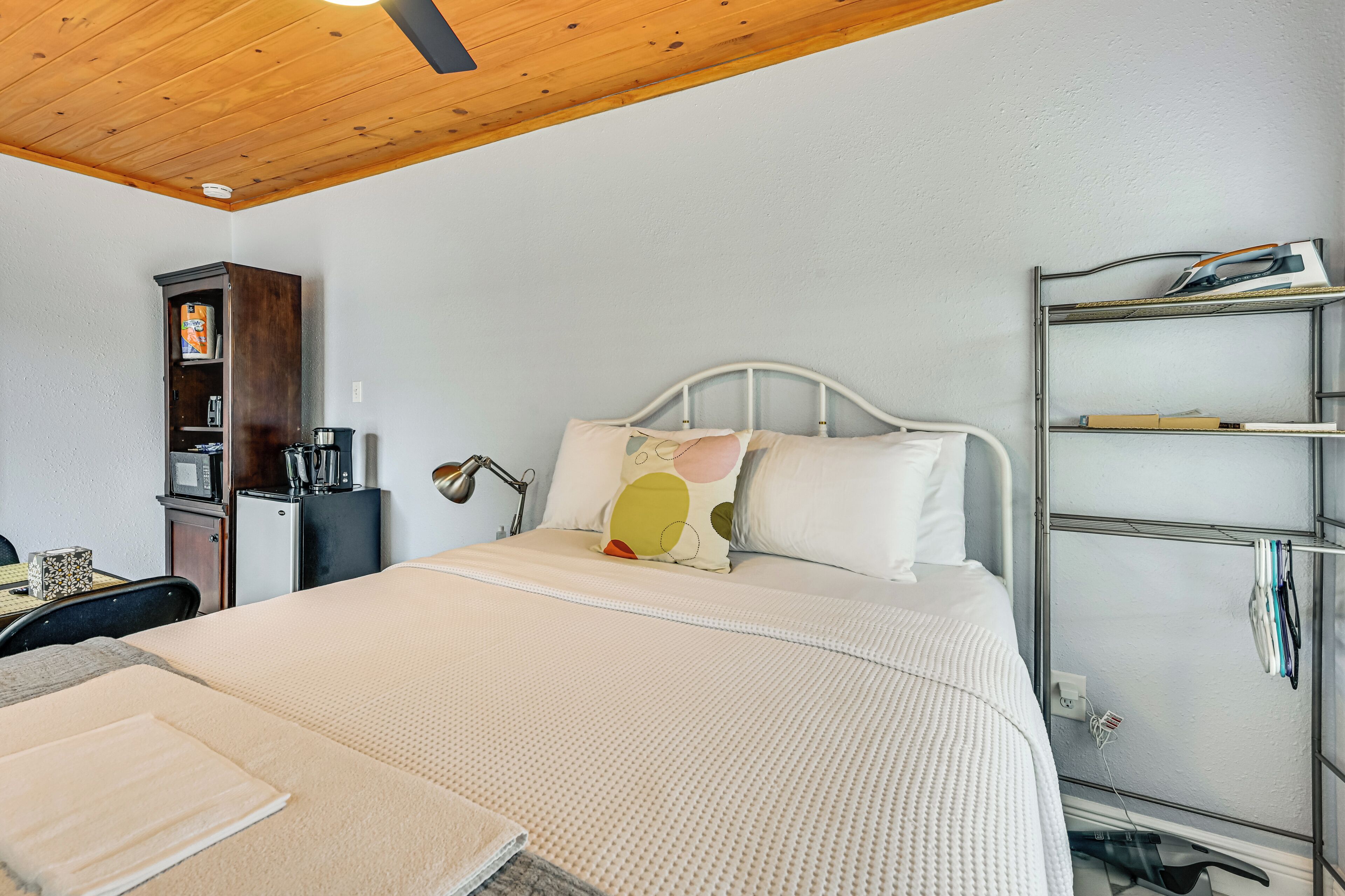 A bedroom with a bed, a desk, a shelf with books, a microwave, and a refrigerator.