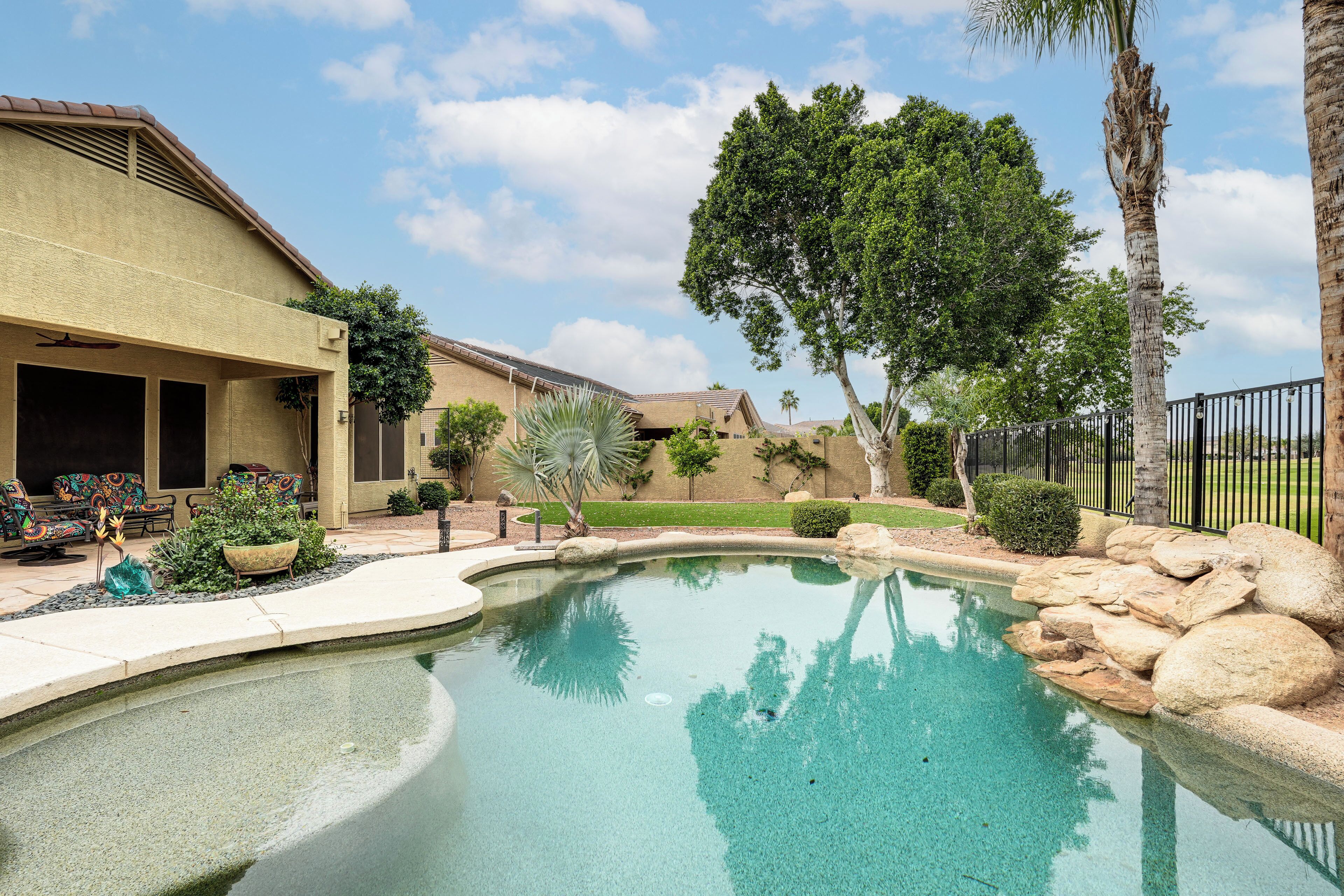 Une piscine à bord incurvé, entourée de rochers et de plantes, avec une maison et du mobilier de patio en arrière-plan.