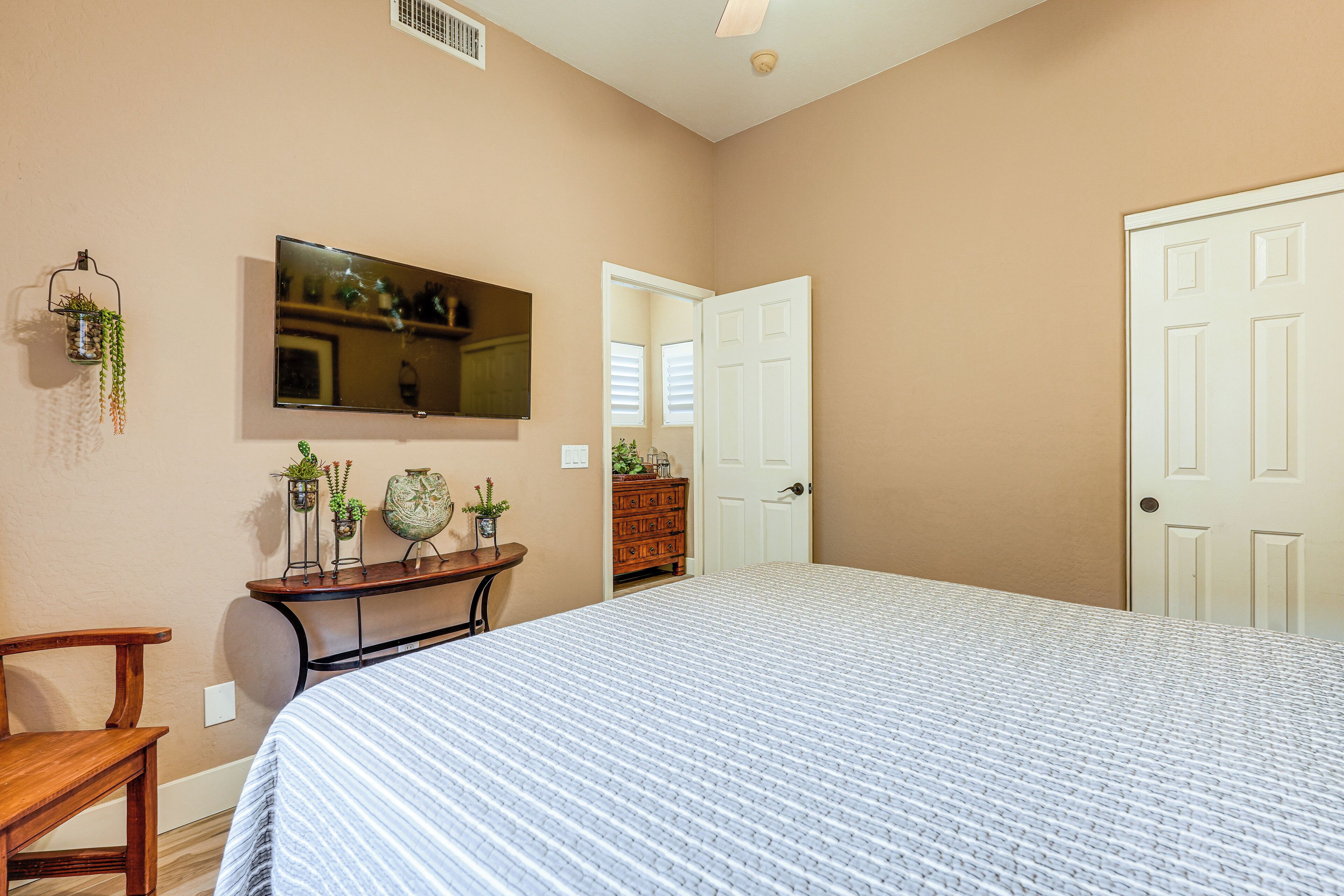 A bedroom with a large bed, a TV mounted on the wall, a wooden dresser, and a door leading to another room.