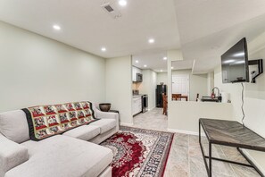 A living room with a sofa, a coffee table, a red and white patterned rug, a kitchen in the background, and a TV mounted on the wall.