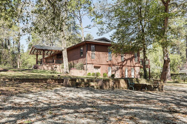 A rustic log cabin with a porch, surrounded by trees and a gravel path.