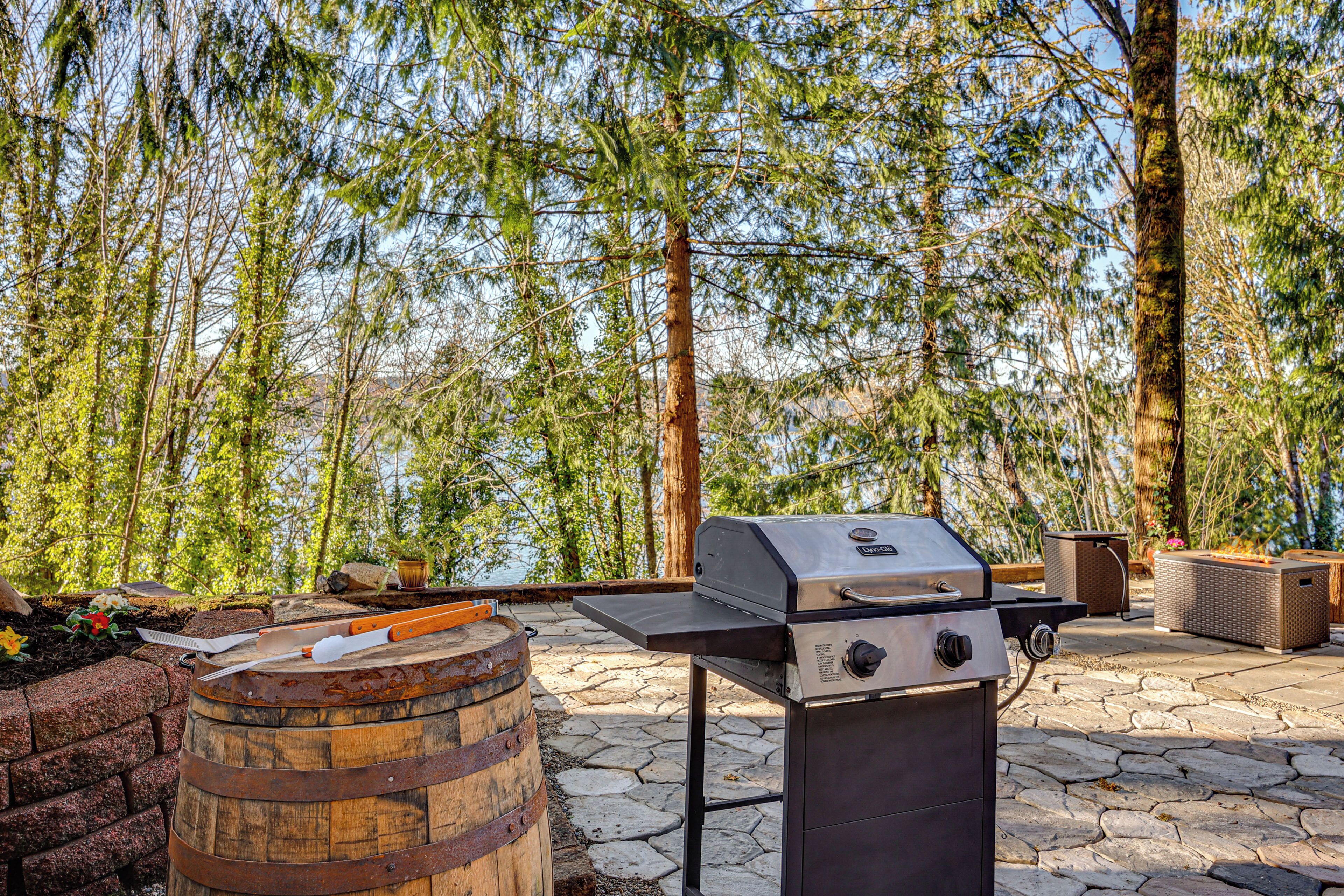 An outdoor grill with a stone patio, surrounded by trees and a view of a body of water.