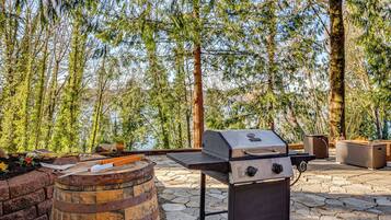 An outdoor grill with a stone patio, surrounded by trees and a view of a body of water.