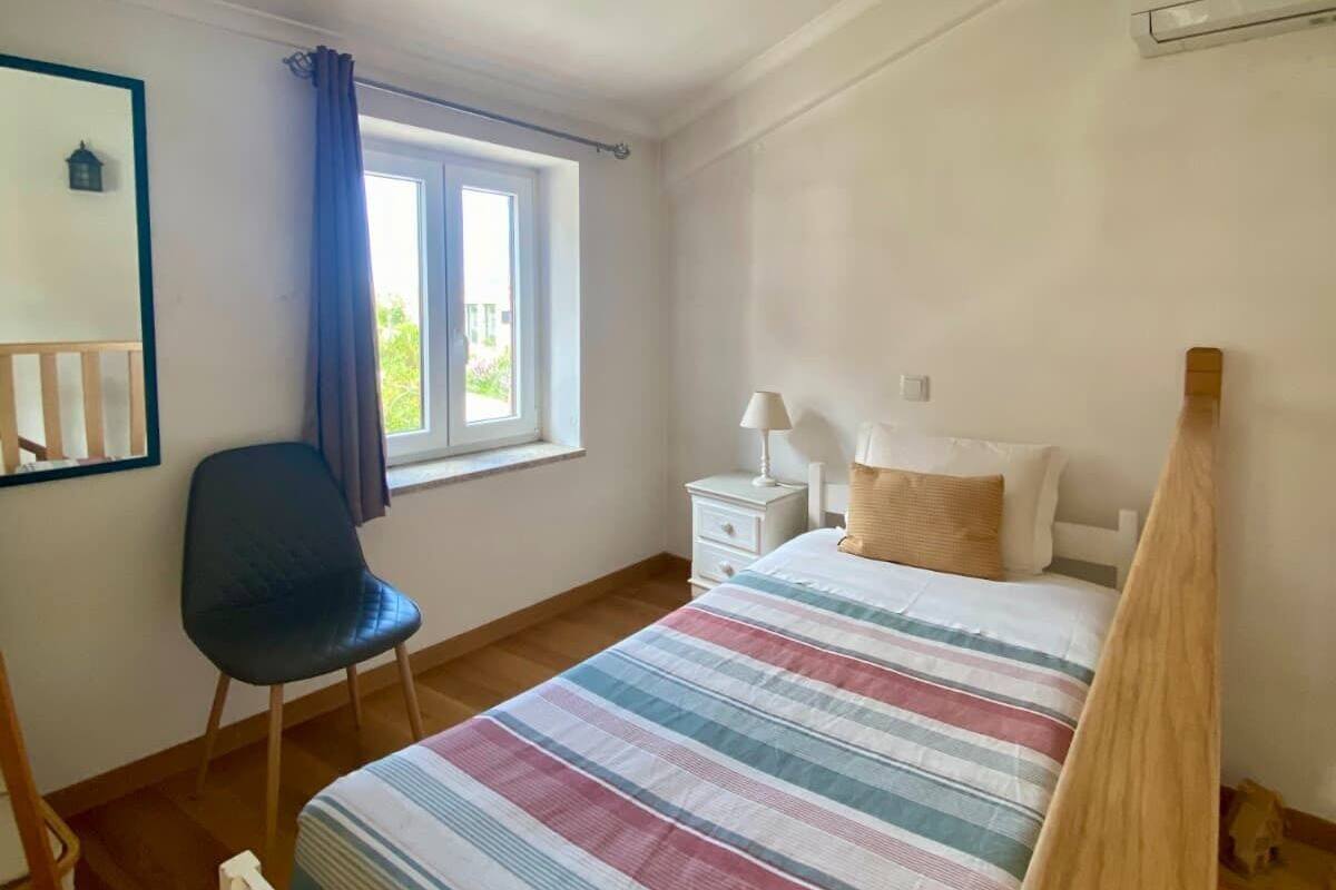 A bedroom with a bed, a chair, a window with curtains, a nightstand, and a wall-mounted air conditioner.