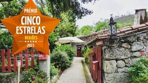 A stone building with a pathway leading to a door, surrounded by greenery and a red fence. - Casas Da Ribeira (Seia)