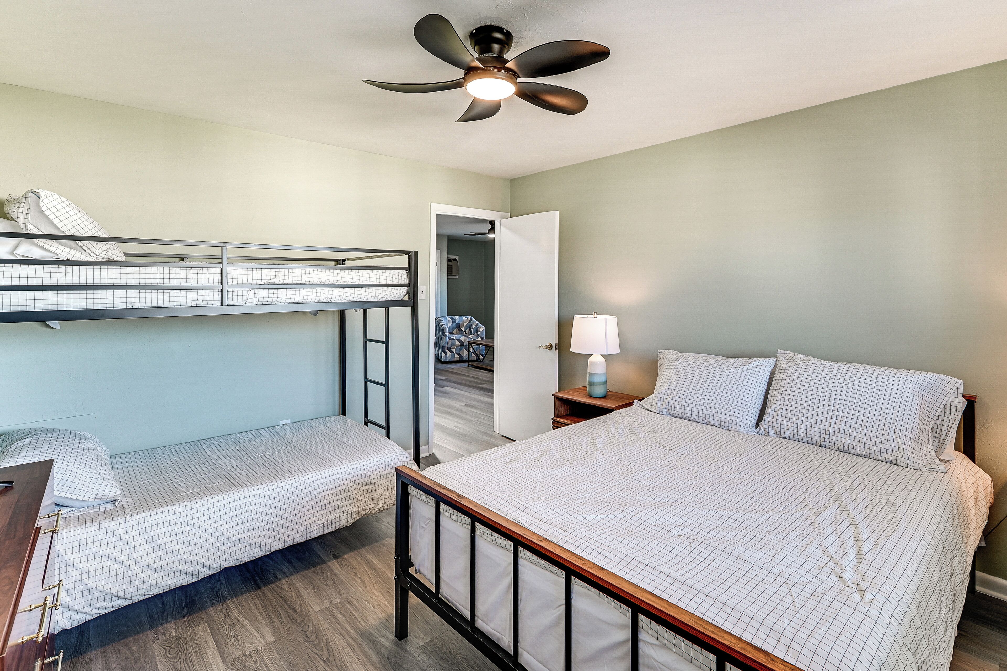 A bedroom with a bunk bed, a single bed, a ceiling fan, a nightstand with a lamp, and a door leading to another room.