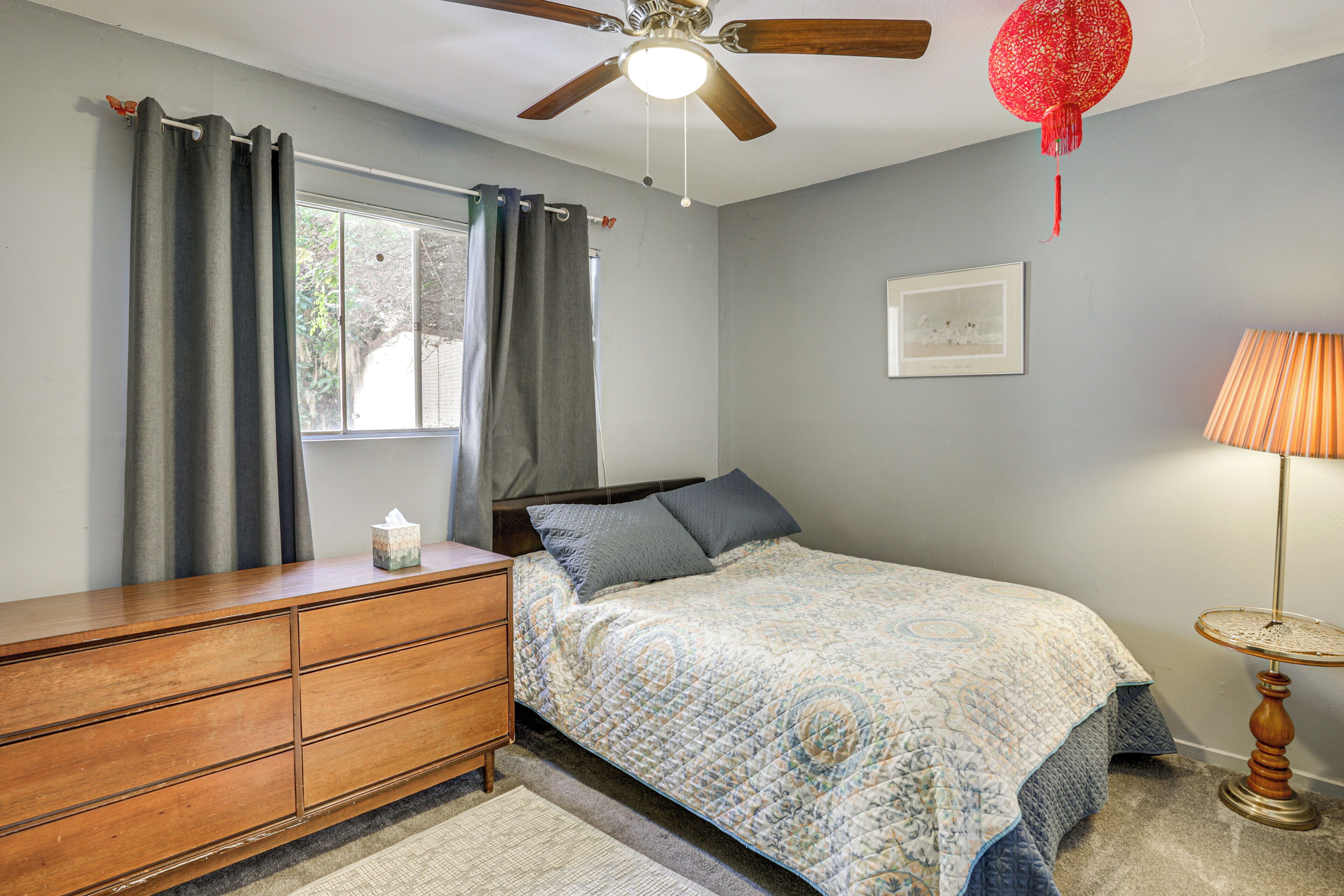 A bedroom with a bed, a dresser, a ceiling fan, a window with curtains, and a lamp.