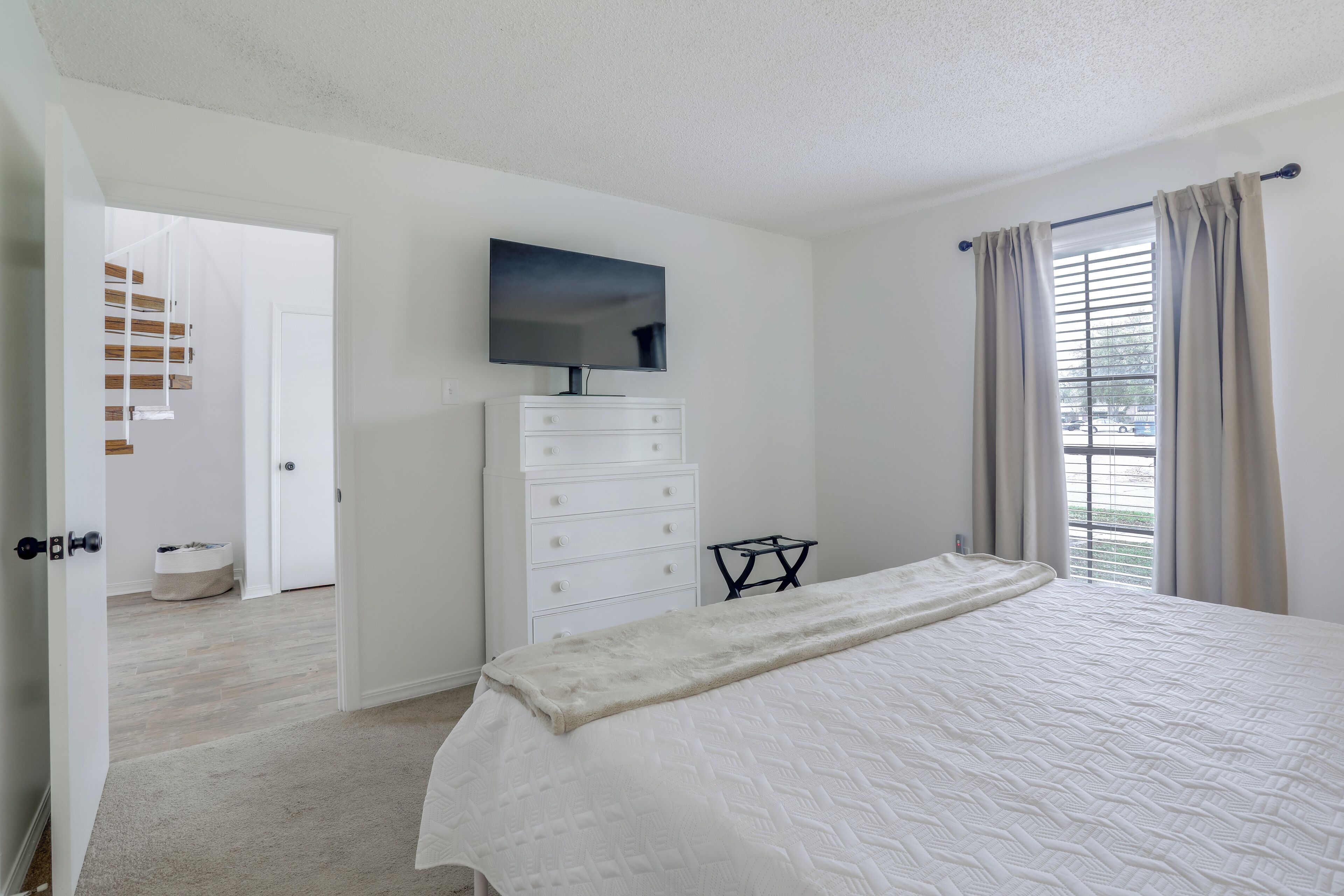 A bedroom with a bed, a television on the wall, a dresser, and a window with curtains.