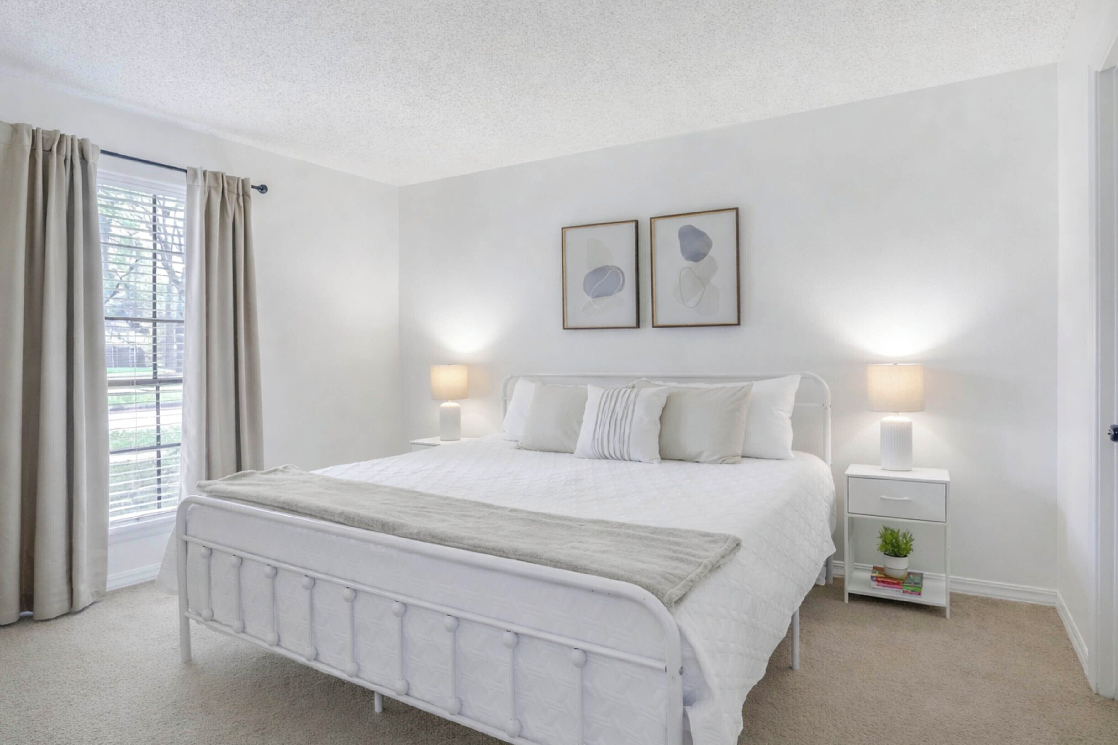 A bedroom with a white bed, bedside tables, a window with curtains, and two framed pictures on the wall.