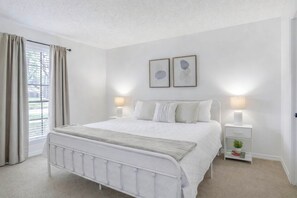Un dormitorio con una cama blanca, mesitas de noche, un ventanal con cortinas y dos cuadros enmarcados en la pared.