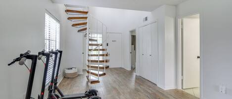 Un hall d’entrée spacieux avec un escalier en bois, des murs blancs et une rangée de trottinettes électriques.