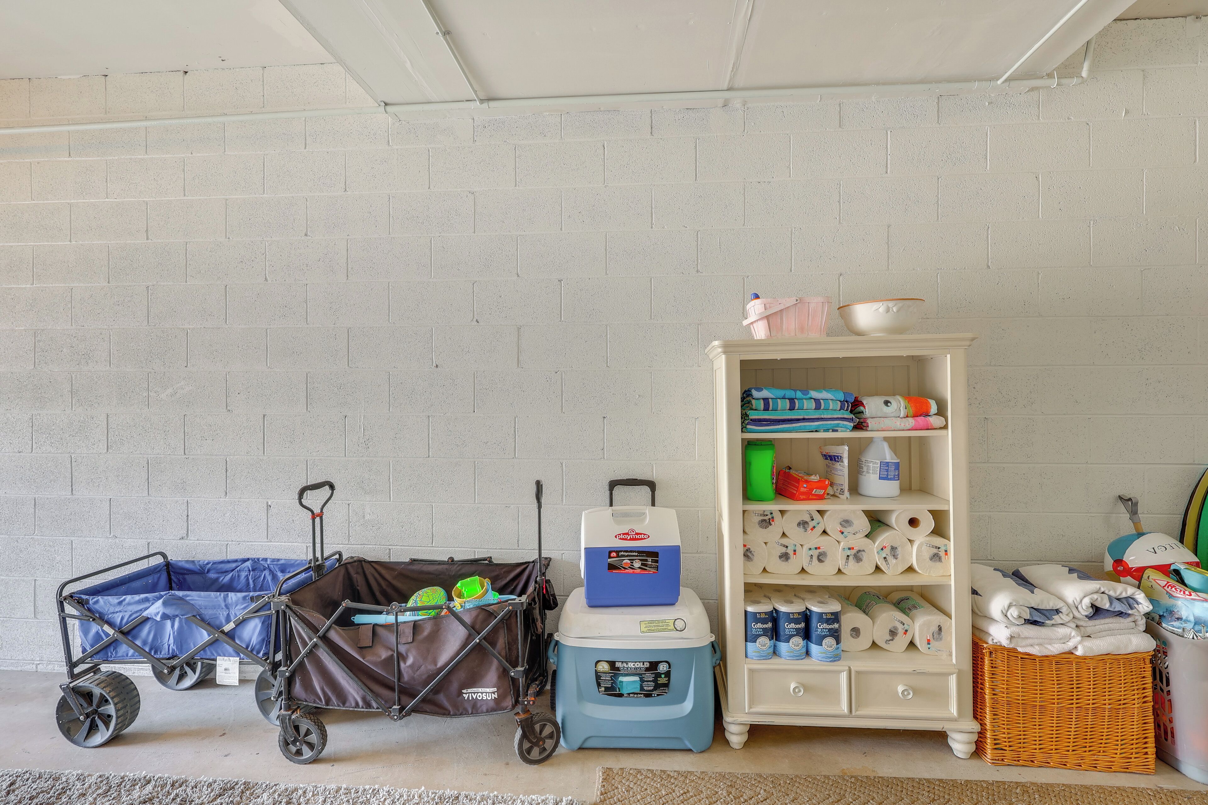 Quarto com parede de tijolos brancos, carrinho de bebê, caixa térmica, cesta de vime e prateleira com artigos para bebês.