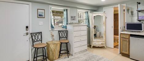 A room with a white dresser, two chairs, a small wooden table, a mirror, a television, and a door leading to another room.