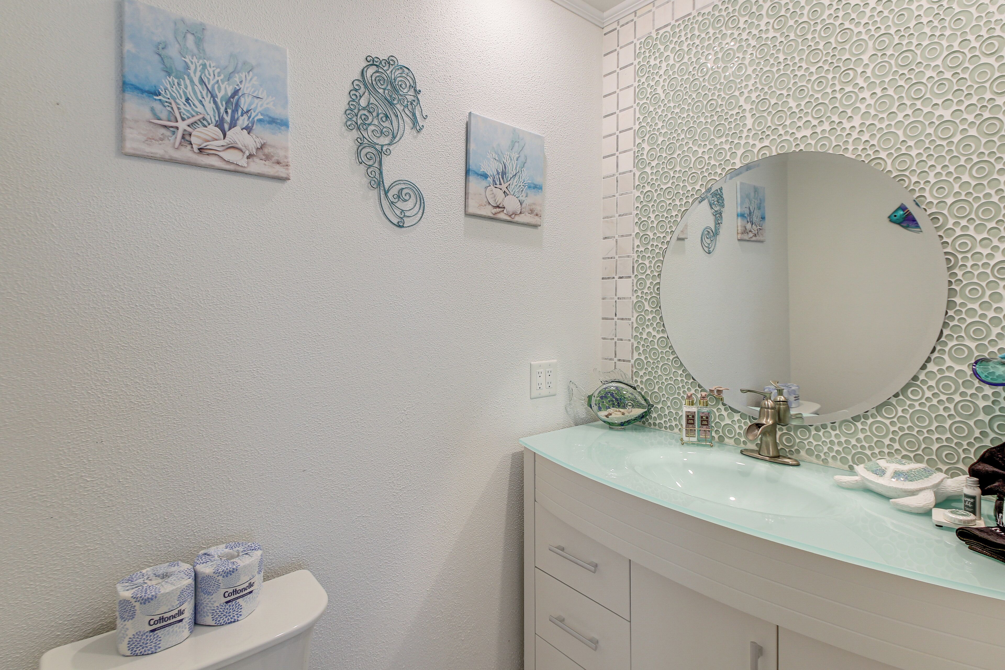 A bathroom with a white sink, a glass countertop, a round mirror, and decorative wall art.