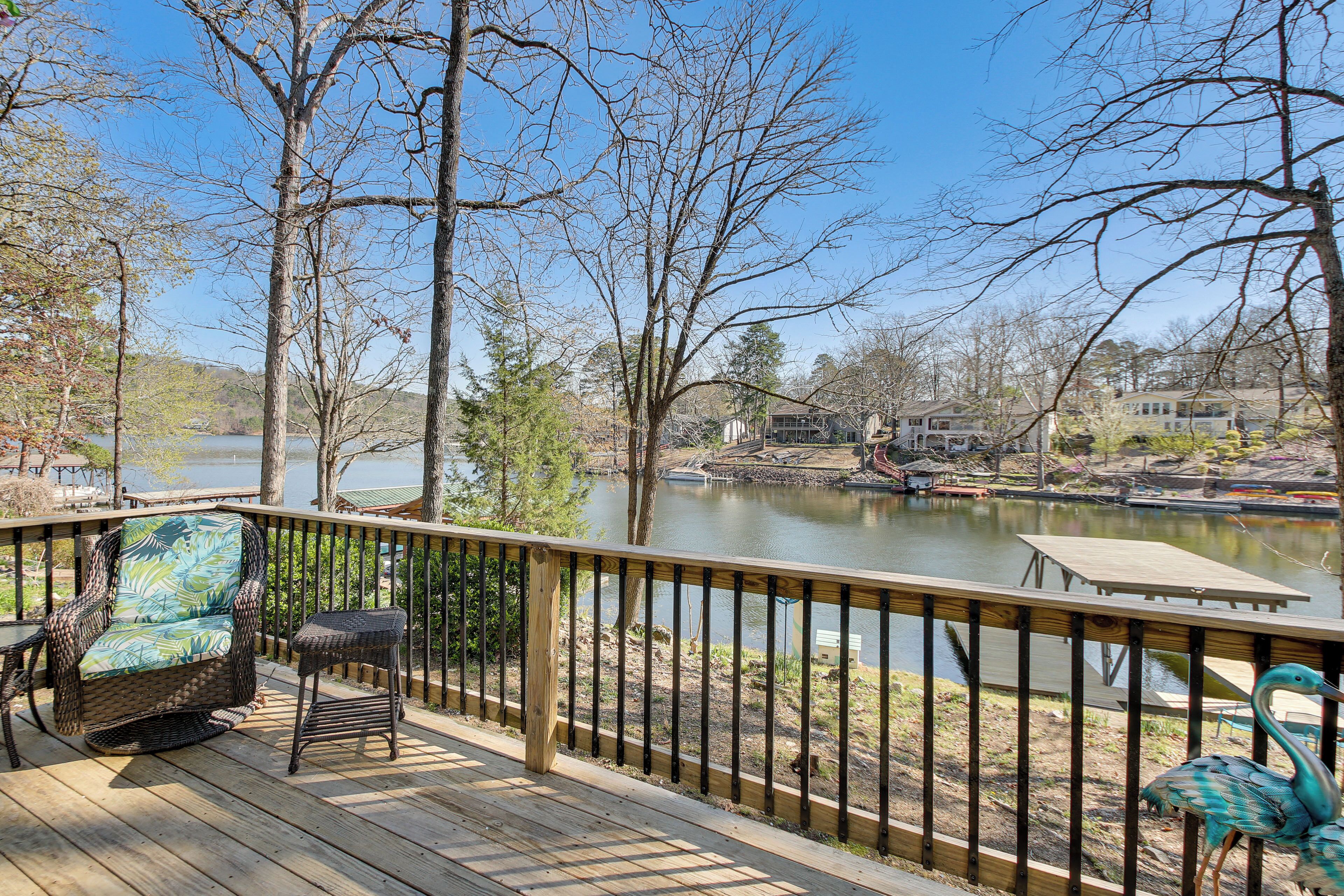 A wooden deck with wicker furniture overlooking a lake, surrounded by trees and houses.