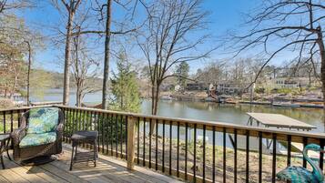 A wooden deck with wicker furniture overlooking a lake, surrounded by trees and houses.