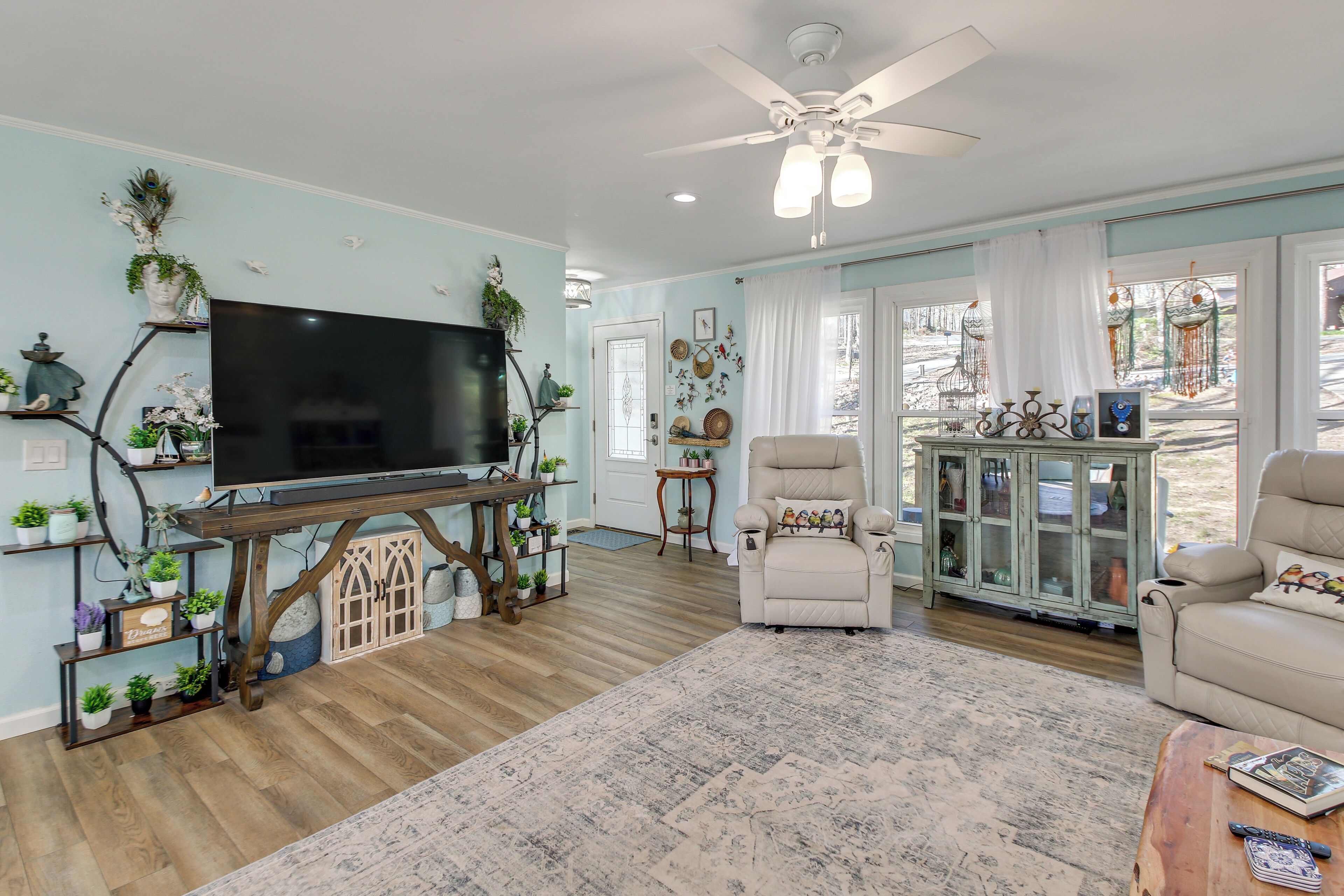 A living room with a TV, a ceiling fan, a beige armchair, a wooden coffee table, and a large window with curtains.