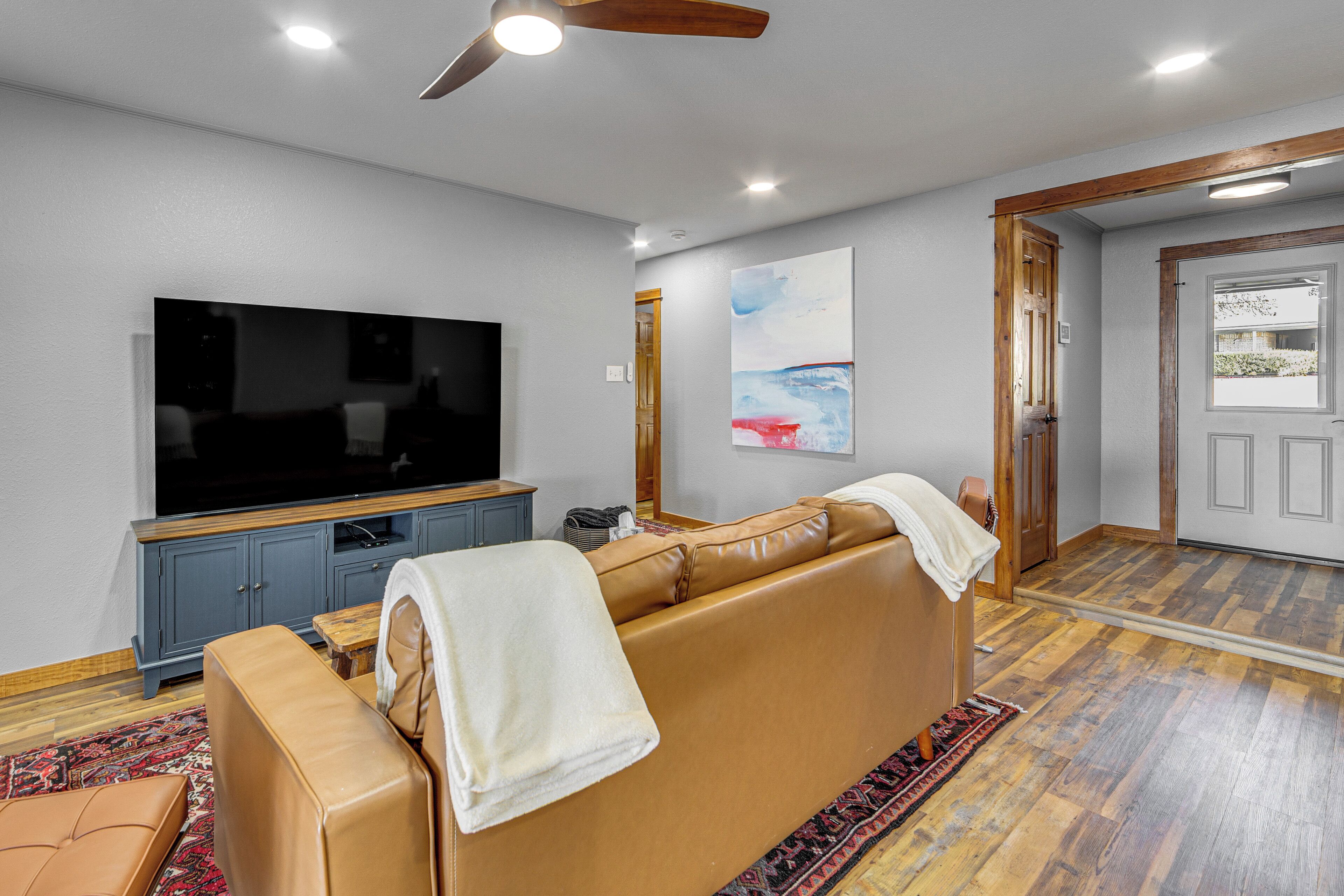 A living room with a large flat-screen TV, a sofa, a wooden ceiling fan, and a door leading to another room.