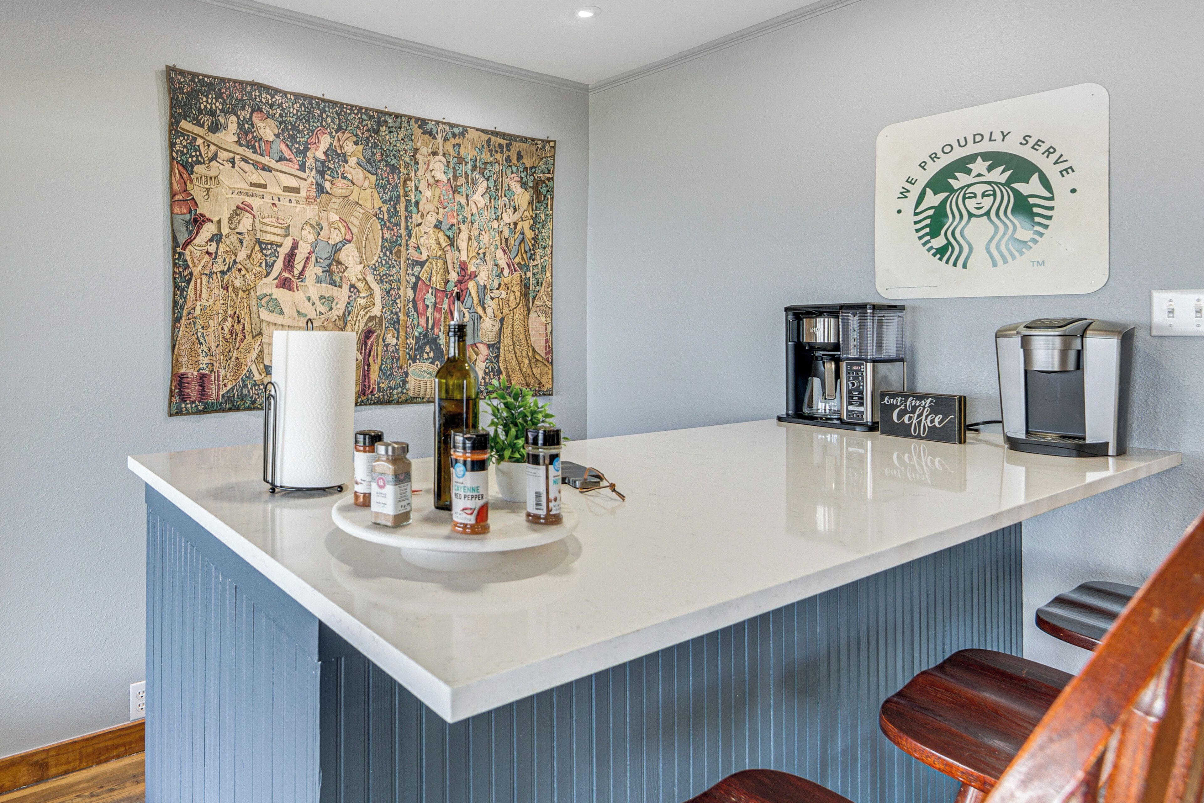 A kitchen with a central island, a coffee machine, and a large tapestry on the wall.
