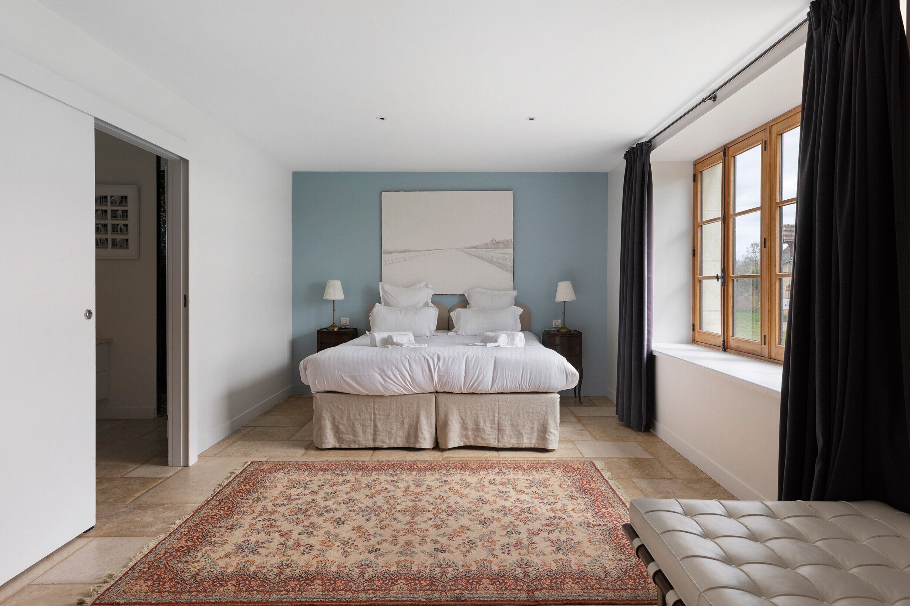 Quarto com cama, abajures, uma janela grande com cortinas, um tapete e uma porta que dá para outro cômodo.