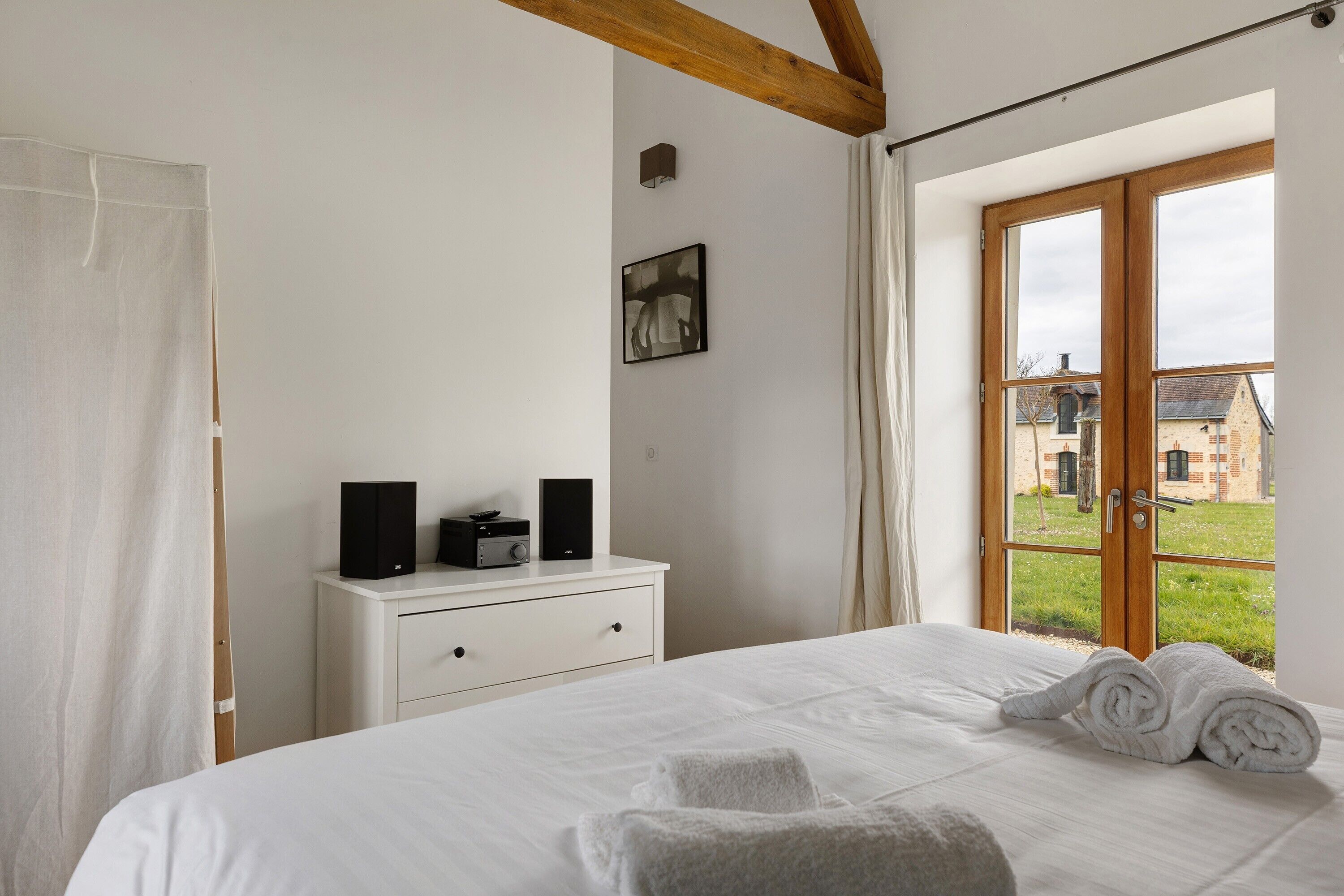 Un dormitorio con cama, una mesita de noche con altavoces, una ventana con cortinas y vistas a edificios.