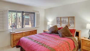 A bedroom with a bed, a dresser, a nightstand, a window with blinds, and a wall-mounted lamp.