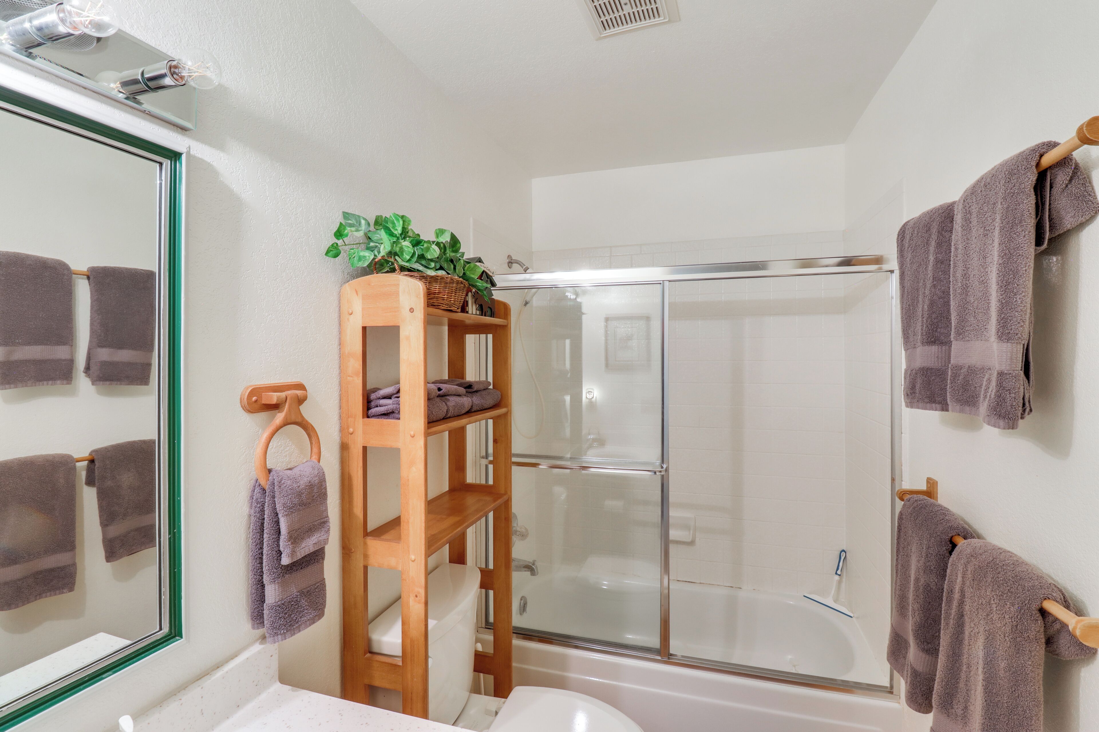 A bathroom with a shower, a wooden shelf with towels, a mirror, and a plant.