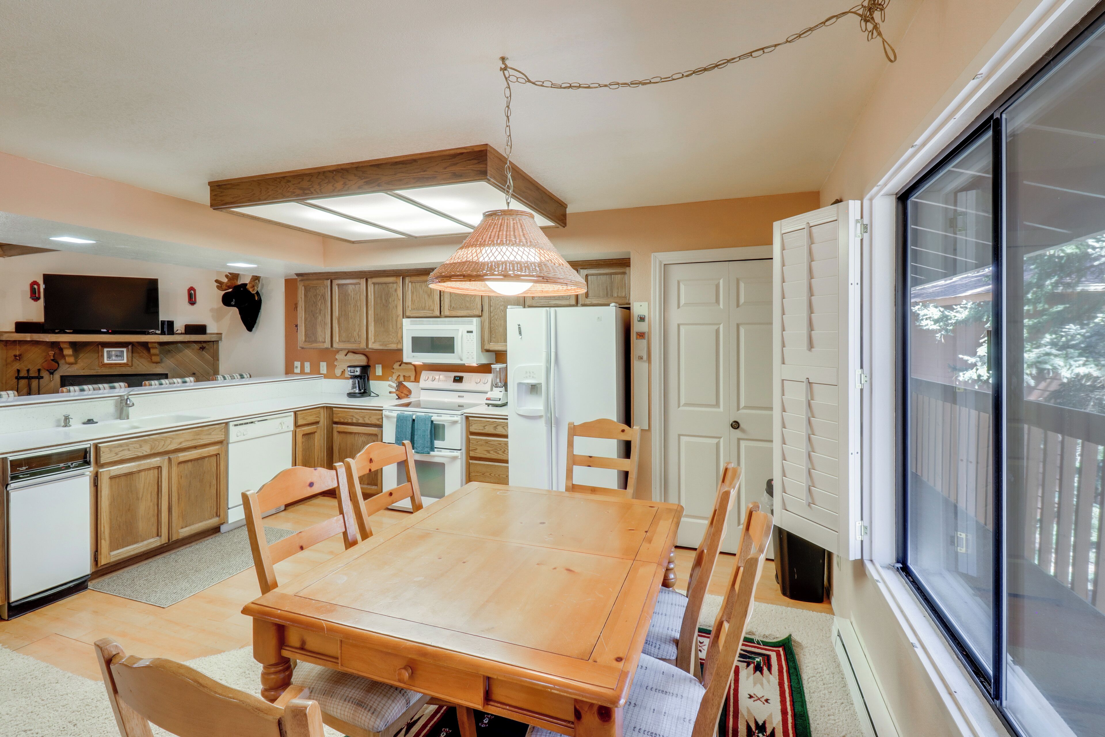 Une cuisine avec des meubles de rangement en bois, une table à manger, des chaises, un luminaire suspendu, un four à micro-ondes, un réfrigérateur et une grande fenêtre.