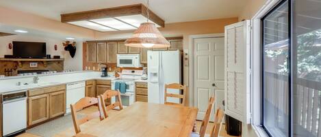 Une cuisine avec des meubles de rangement en bois, une table à manger, des chaises, un luminaire suspendu, un four à micro-ondes, un réfrigérateur et une grande fenêtre.
