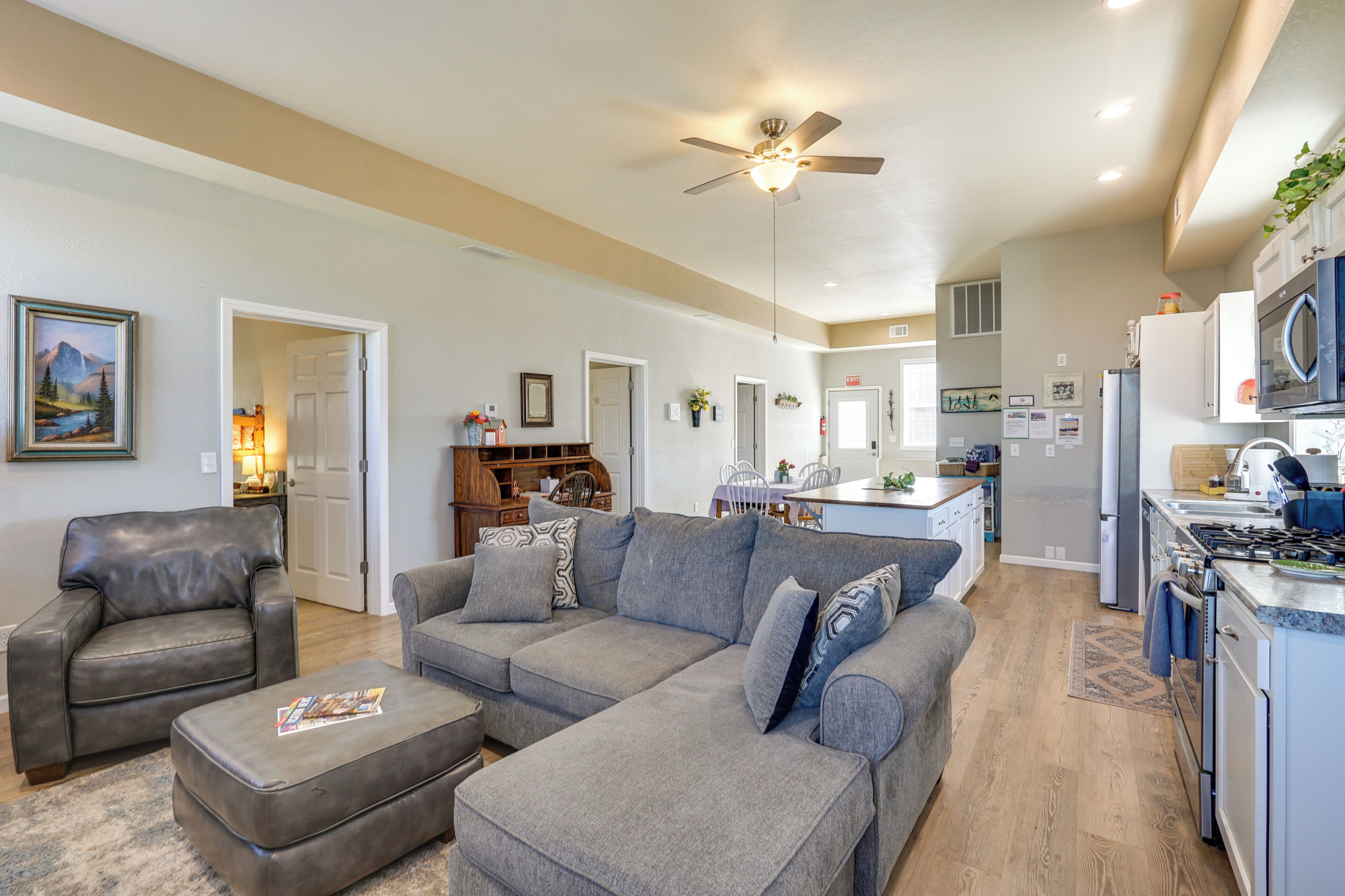 A modern kitchen with a microwave, stove, and sink, a gray sectional sofa, a leather armchair, and a ceiling fan.