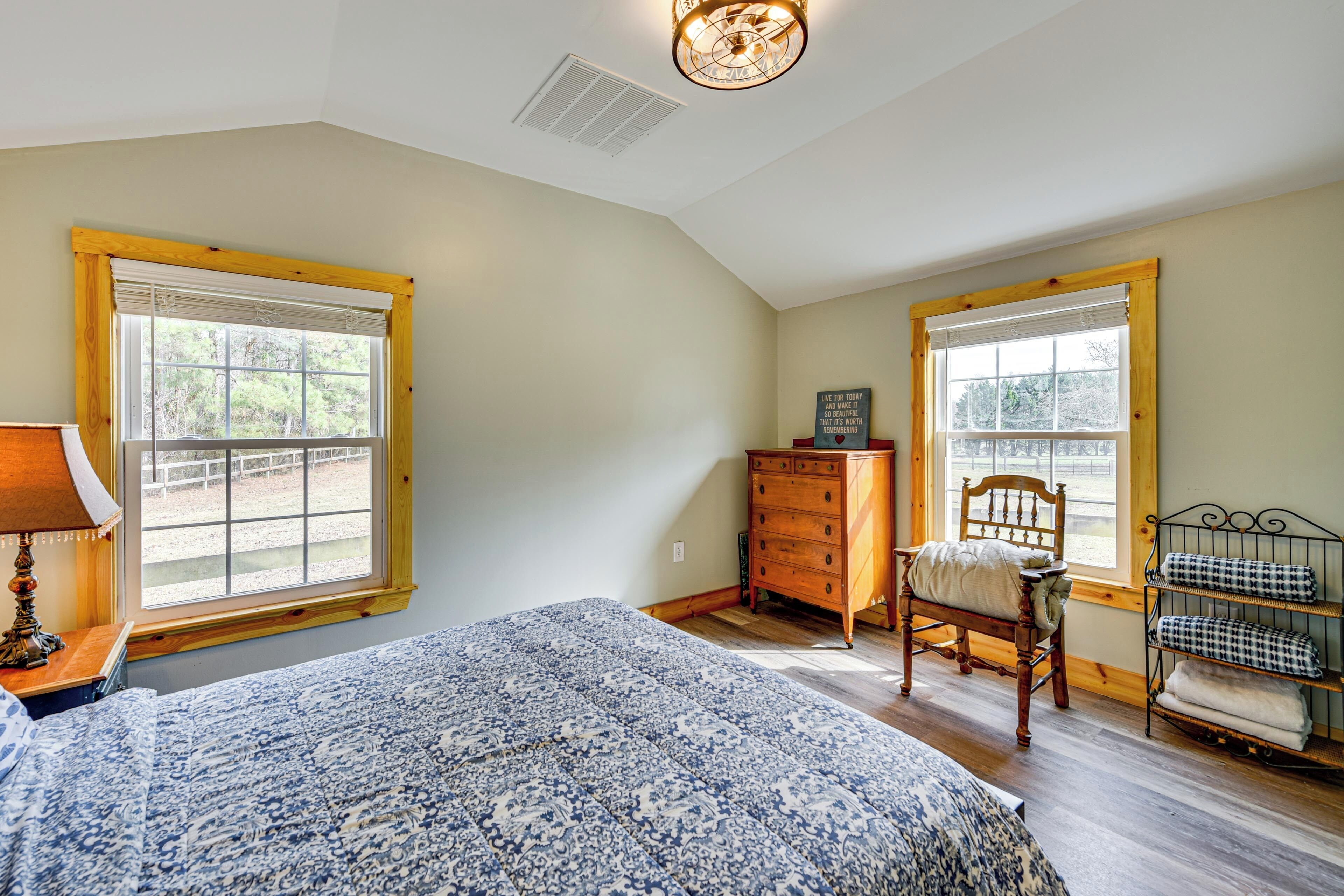 A bedroom with a large bed, two windows, a dresser, a chair, and a metal shelf with folded blankets.