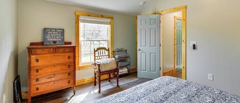 A bedroom with a wooden dresser, a bed with a patterned bedspread, a chair, a window with blinds, and a door.