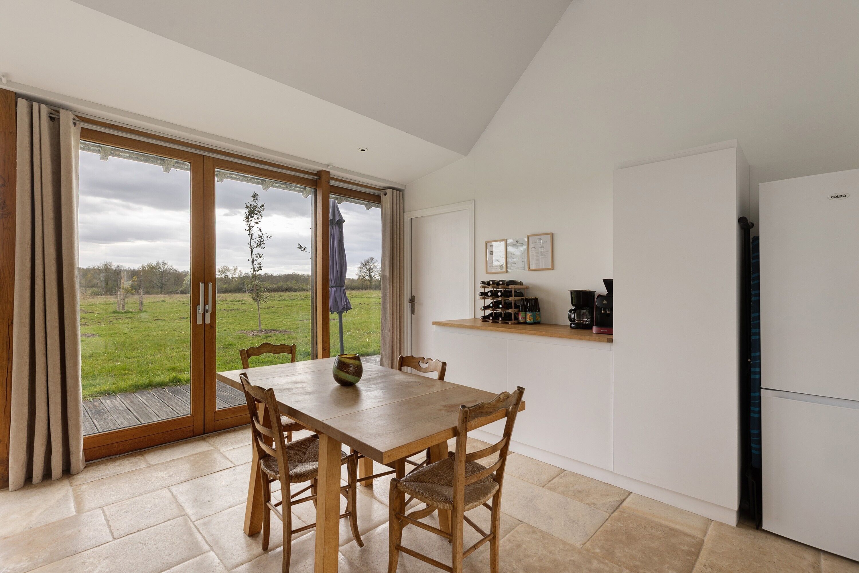 Een eetgedeelte met een tafel en stoelen, een keuken met apparatuur, en uitzicht op een grasveld door grote glazen deuren.
