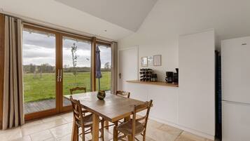 Een eetgedeelte met een tafel en stoelen, een keuken met apparatuur, en uitzicht op een grasveld door grote glazen deuren.