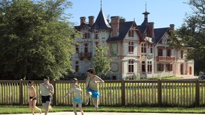 A swimming pool with a wooden fence in the foreground and a large, historic building with multiple chimneys and a tower in the background.